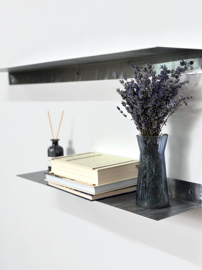 May include: Two horizontal metal wall shelves with a brushed finish. The lower shelf holds a stack of three books, a small dark glass bottle with reed diffuser sticks, and a ribbed blue glass vase filled with a bouquet of dried purple lavender.