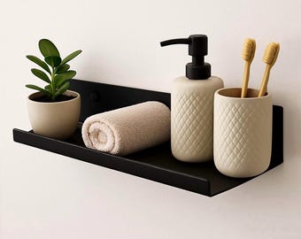Puede incluir: Una estantería de metal negro sostiene una pequeña planta en maceta, una toalla beige enrollada, un dispensador de jabón crema con una bomba negra y un portacepillos crema con dos cepillos de dientes de bambú. La estantería está montada en una pared blanca.