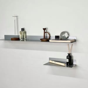 Puede incluir: Dos estantes de pared de metal gris mate montados horizontalmente. El estante superior sostiene un soporte para incienso fino, una botella de vidrio transparente, un candelabro de madera oscura, una caja negra peque&ntilde;a y una esfera de vidrio. El estante inferior sostiene un tubo blanco y una botella negra.