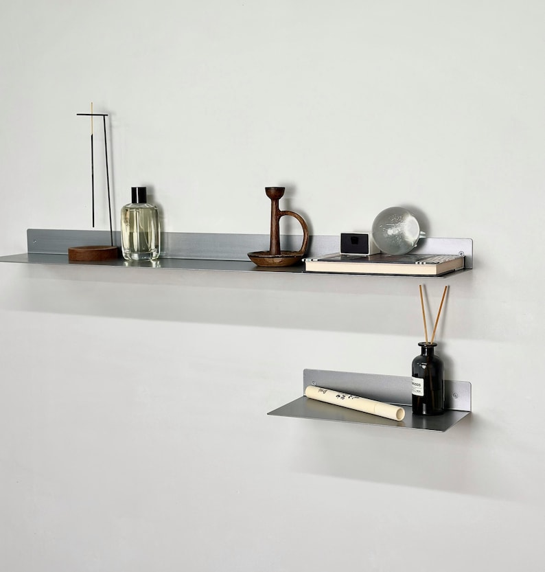 Puede incluir: Dos estantes de pared de metal gris mate montados horizontalmente. El estante superior sostiene un soporte para incienso fino, una botella de vidrio transparente, un candelabro de madera oscura, una caja negra peque&ntilde;a y una esfera de vidrio. El estante inferior sostiene un tubo blanco y una botella negra.