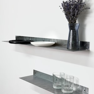 May include: Two metal wall shelves with items on them. The top shelf holds a black plate, a white plate, and a blue vase with dried lavender. The bottom shelf has two clear drinking glasses.