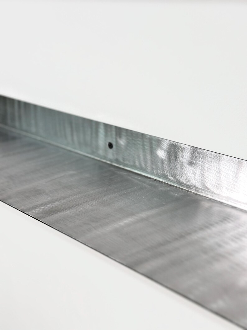 May include: A long, rectangular strip of galvanized steel with a brushed metallic finish. The metal piece features a vertical back edge bent at a ninety-degree angle, and a single, small, circular pre-drilled hole centered along the back flange.