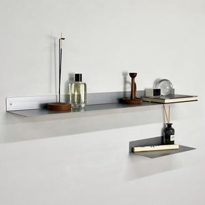 Puede incluir: Dos estantes de pared de metal gris mate montados a diferentes alturas. El estante superior, m&aacute;s largo, sostiene un porta-incienso, un frasco de perfume de vidrio, un portavelas de madera, una peque&ntilde;a caja blanca y una esfera de vidrio. El estante inferior, m&aacute;s corto, sostiene un frasco de vidrio y un tubo blanco.