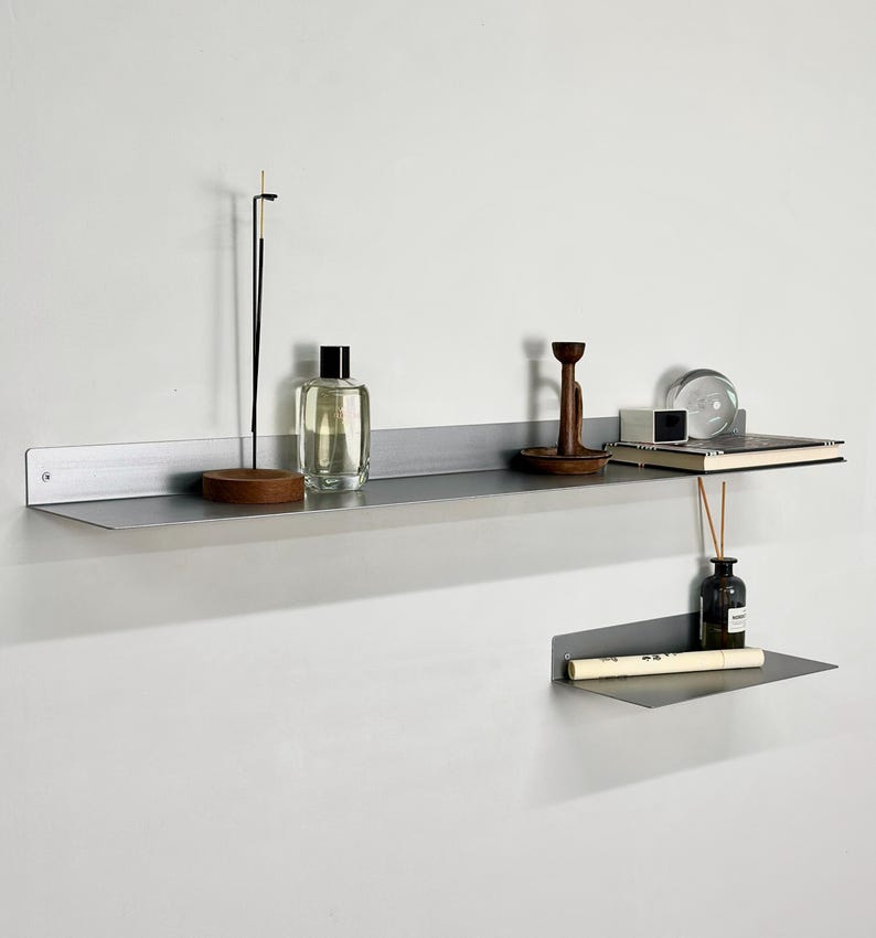 Puede incluir: Dos estantes de pared de metal gris mate montados a diferentes alturas. El estante superior, m&aacute;s largo, sostiene un porta-incienso, un frasco de perfume de vidrio, un portavelas de madera, una peque&ntilde;a caja blanca y una esfera de vidrio. El estante inferior, m&aacute;s corto, sostiene un frasco de vidrio y un tubo blanco.
