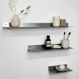 May include: Three horizontal metal wall shelves with a dark, brushed finish and pre-drilled mounting holes. The shelves hold a textured vase with dried stems, a ceramic cup, a bar of soap, a dark bottle, a tube, toothbrushes, and a small bowl.
