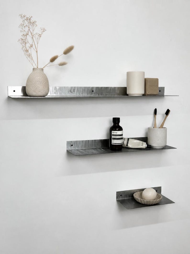 May include: Three horizontal metal wall shelves with a dark, brushed finish and pre-drilled mounting holes. The shelves hold a textured vase with dried stems, a ceramic cup, a bar of soap, a dark bottle, a tube, toothbrushes, and a small bowl.