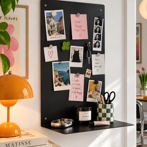 May include: A rectangular black metal wall-mounted magnetic memo board with a small integrated shelf at the bottom. The board displays various photographs, notes with handwritten text, a set of keys, a candle, and a green and white checkered cup.
