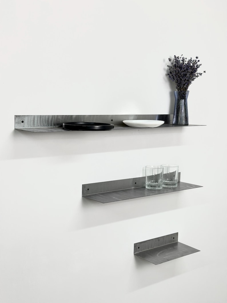 May include: Three wall-mounted shelves made of dark gray metal with a visible brushed texture. The shelves feature a flat horizontal surface and a vertical back panel with pre-drilled holes for mounting, arranged in a staggered vertical formation.