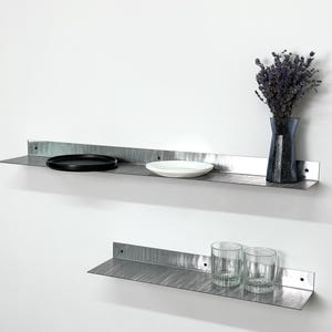 May include: Two horizontal brushed metal wall shelves with an L-shaped profile. The top shelf holds a black circular plate, a white bowl, and a dark blue glass vase with dried flowers. The bottom shelf holds two clear glass tumblers.