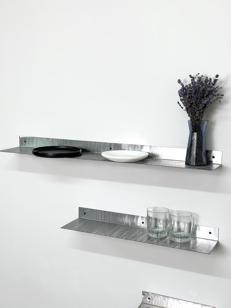 May include: Two horizontal brushed metal wall shelves with an L-shaped profile. The top shelf holds a black circular plate, a white bowl, and a dark blue glass vase with dried flowers. The bottom shelf holds two clear glass tumblers.