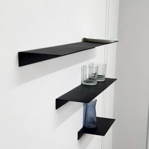 Puede incluir: Tres estantes de pared de metal negro mate montados en disposici&oacute;n escalonada. El estante superior sostiene un peque&ntilde;o plato rectangular blanco. El estante central sostiene dos vasos de cristal transparente. El inferior sostiene un jarr&oacute;n azul estriado.