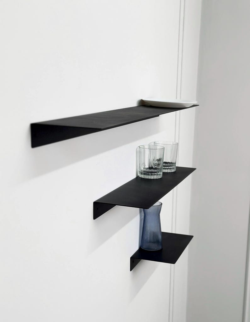 Puede incluir: Tres estantes de pared de metal negro mate montados en disposici&oacute;n escalonada. El estante superior sostiene un peque&ntilde;o plato rectangular blanco. El estante central sostiene dos vasos de cristal transparente. El inferior sostiene un jarr&oacute;n azul estriado.