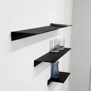 May include: Three black metal floating shelves mounted on a white wall. The top shelf holds a white plate. The middle shelf has two clear drinking glasses. The bottom shelf displays a blue vase.