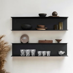 May include: Three-tiered black metal shelves against a white wall, displaying various home decor items. The shelves hold bowls, plates, glasses, and decorative objects in neutral tones, creating a minimalist aesthetic. The items include a variety of textures and shapes.