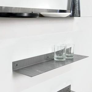 May include: Three stainless steel floating shelves mounted on a white wall. The top shelf holds a black plate, a white plate, and a blue glass vase with lavender. The middle shelf has two clear drinking glasses. The bottom shelf is empty.