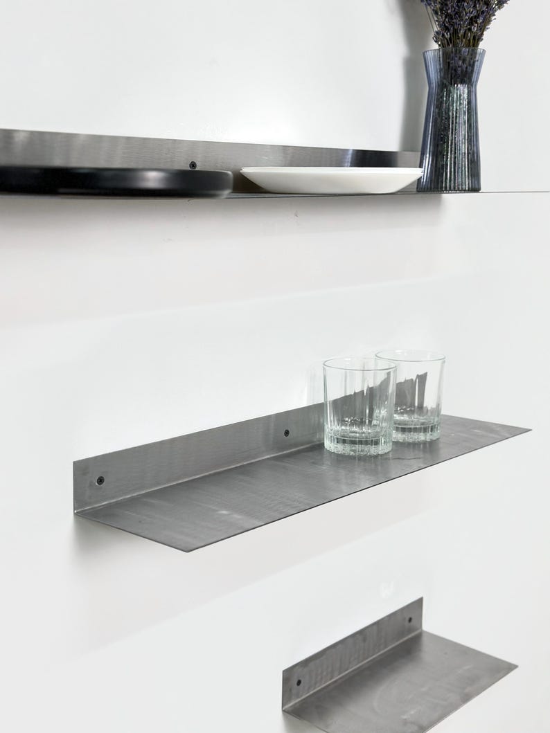May include: Three stainless steel floating shelves mounted on a white wall. The top shelf holds a black plate, a white plate, and a blue glass vase with lavender. The middle shelf has two clear drinking glasses. The bottom shelf is empty.