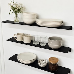 Pode incluir: Três prateleiras flutuantes de metal preto fosco montadas horizontalmente. A prateleira superior contém um vaso de vidro com flores brancas, uma pilha de três tigelas creme e quatro pratos ovais. A prateleira do meio apresenta duas canecas, dois copos estriados e três tigelas cinzas. A prateleira inferior exibe uma pilha de pratos, uma pequena tigela estampada e um pote de vidro com tampa de madeira.