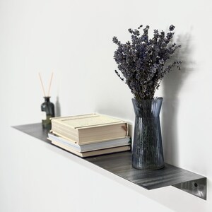 May include: A dark gray metal wall shelf holding a stack of two beige books, a clear ribbed glass vase filled with dried purple lavender stems, and a small dark glass bottle with thin wooden diffuser reeds protruding from the top.