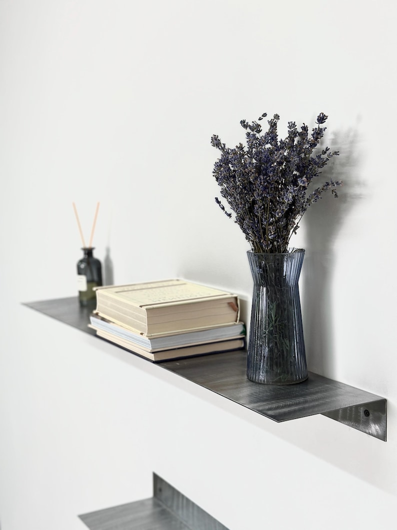 May include: A dark gray metal wall shelf holding a stack of two beige books, a clear ribbed glass vase filled with dried purple lavender stems, and a small dark glass bottle with thin wooden diffuser reeds protruding from the top.