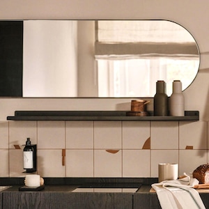 Puede incluir: Escena de cocina moderna con un gran espejo ovalado con marco negro sobre una estantería oscura. La estantería contiene botellas decorativas y un cuenco de madera. Debajo, un dispensador de jabón y una vela están en la encimera, con un salpicadero de azulejos.