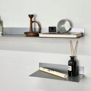 Puede incluir: Dos estantes flotantes de metal plateado montados en una pared blanca. El estante superior contiene un candelabro de madera, una caja negra, un libro y una esfera de cristal. El estante inferior muestra un pergamino y una botella de vidrio oscuro con cañas.