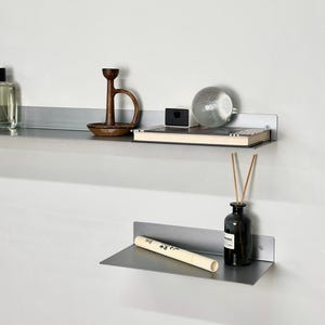 Puede incluir: Dos estantes horizontales montados en la pared hechos de metal gris cepillado. El estante superior sostiene una botella de vidrio transparente, un portavelas de madera marr&oacute;n, una caja negra peque&ntilde;a y un libro. El estante inferior sostiene un tubo crema.