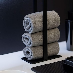 Pode incluir: Um moderno suporte de toalhas em metal preto com três toalhas cinzentas enroladas. O suporte tem um design minimalista e está colocado sobre uma superfície branca, com um fundo preto. As toalhas parecem macias e fofas.