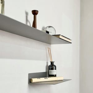 Puede incluir: Dos estantes de pared de metal gris mate montados uno sobre otro. El estante superior sostiene un portavelas de madera oscura, una esfera de vidrio transparente y un libro. El estante inferior sostiene una botella de vidrio oscuro y un tubo.