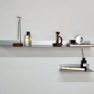 May include: A minimalist display featuring two metal shelves mounted on a light gray wall. The top shelf holds a wooden incense holder with incense sticks, a perfume bottle, and a wooden candle holder. The bottom shelf displays a scroll and a small bottle.