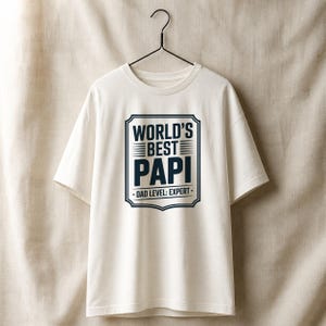 May include: A white t-shirt with the text "WORLD'S BEST PAPI" in a navy blue framed design. Below the text is "DAD LEVEL: EXPERT" in a smaller font. The shirt is hanging on a black hanger.