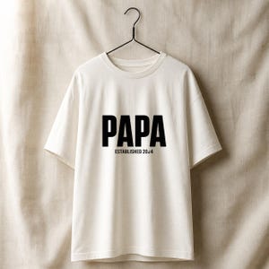 Könnte beinhalten: Ein weißes T-Shirt mit dem Wort "PAPA" in fetten schwarzen Buchstaben und "ESTABLISHED 2024" darunter. Das T-Shirt hängt an einem schwarzen Kleiderbügel vor einem neutralen Hintergrund. Das T-Shirt hat einen Rundhalsausschnitt und kurze Ärmel.