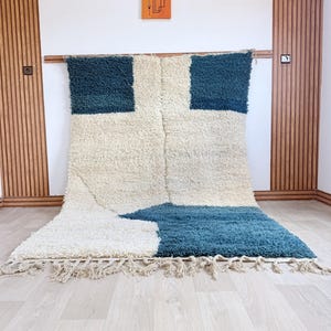 Peut inclure: Un grand tapis rectangulaire avec un motif géométrique. Le tapis présente un fond crème avec deux carrés bleu foncé et une section bleue en bas. Le tapis a une texture touffue et une frange sur le bord inférieur.