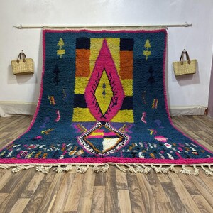 Può includere: Un grande tappeto rettangolare con un vivace design geometrico. Il tappeto presenta uno sfondo verde acqua con una forma a diamante rosa e giallo centrale. Il bordo è rosa e il tappeto ha frange color crema.