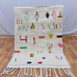 Puede incluir: Alfombra rectangular de color crema con diseños geométricos coloridos. La alfombra presenta una superficie texturizada y una franja en el borde inferior. El diseño incluye formas en verde, naranja, rosa, amarillo y negro.