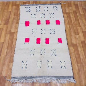 May include: A rectangular, off-white rug with a geometric pattern. The rug features a series of dark blue and hot pink shapes arranged across the surface. The edges are finished with a dark blue border and fringe.