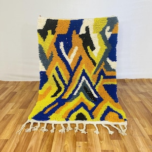 May include: A colorful, geometric patterned rug with a white fringe. The rug features a vibrant design with yellow, blue, orange, black, and white zigzags. The rug is displayed on a wooden floor.