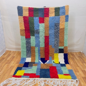 Op de afbeelding: Een kleurrijk, rechthoekig tapijt met een geometrisch patroon. Het tapijt heeft een verscheidenheid aan kleuren, waaronder rood, blauw, groen, geel en bruin. Het heeft een getextureerd oppervlak en witte franjes langs de onderrand. Het tapijt ligt op een houten vloer.