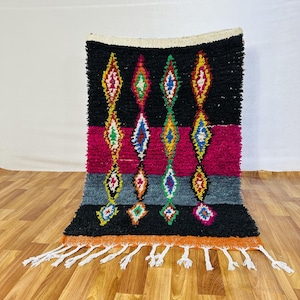 Può includere: Un vivace tappeto tessuto a mano con sfondo nero e motivi a rombi colorati. Il design include strisce rosa, grigie e arancioni, con frange bianche lungo il bordo inferiore. Il tappeto è realizzato in materiale spesso e strutturato.