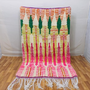 May include: A colorful, rectangular rug with a cream-colored background and a vibrant pattern. The design features vertical lines in shades of pink, orange, and green, with a purple border and white fringe. The rug is made of a thick, woven material.