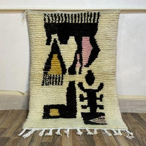 以下が含まれることがあります： 抽象的なデザインのクリーム色の長方形ラグ。 ラグは、テクスチャード加工された背景に黒、ピンク、黄色の幾何学的な形を特徴としています。 白いフリンジが下端を飾り、この床材に装飾的な要素を追加しています。