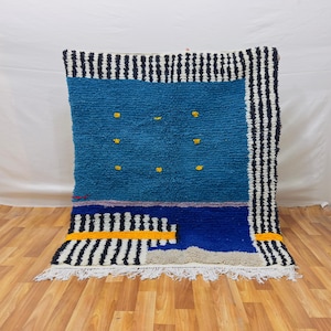 Może przedstawiać: Prostokątny, ręcznie tkany dywan z teksturowanym, niebieskim polem i obramowaniem z czarno-białych pasków. Dywan posiada żółte kropki i geometryczny wzór z żółtym paskiem. Białe frędzle.