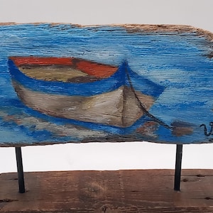 Könnte beinhalten: Ein kleines Holzboot, das in Blau, Weiß und Orange bemalt ist, ist auf einem Holzständer montiert. Das Boot ist auf einem Stück Treibholz mit blauem Hintergrund bemalt. Die Unterschrift des Künstlers, "Vickie", ist in der unteren rechten Ecke sichtbar.