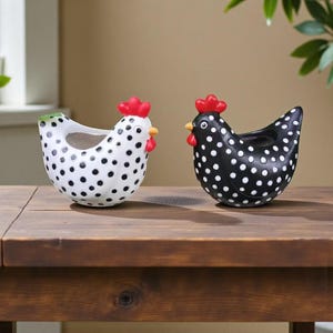 Puede incluir: Dos macetas de cerámica con forma de gallina. Una es blanca con lunares negros, cresta roja y pico amarillo. La otra es negra con lunares blancos, cresta roja y pico amarillo. Ambas están sobre una superficie de madera.
