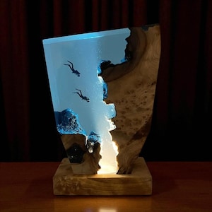 May include: A distinctive wooden lamp with a clear resin section depicting two diver silhouettes, forming an underwater scene. The lamp has a natural wood base and an internal light source, illuminating the scene. The lamp is approximately 30 cm tall.