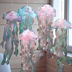 May include: Hanging jellyfish decorations in shades of pink, blue, and green. The jellyfish have a translucent, iridescent appearance with flowing ribbon-like tentacles. Tiny lights are strung throughout the decorations, adding a whimsical touch. The jellyfish are suspended from the ceiling.