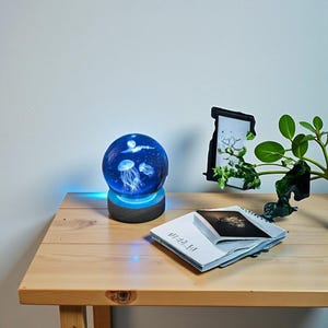Può includere: Lampada decorativa a forma di medusa blu e bianca con base in legno. La lampada ha un globo di vetro sferico con meduse e un design a luna. Un quaderno e una foto incorniciata sono sul tavolo.