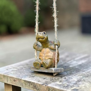 Może przedstawiać: Brązowo-beżowa figurka żółwia siedząca na małej drewnianej huśtawce zawieszonej na dwóch grubych, teksturowanych sznurach jutowych. Żółw ma chropowatą skorupę, widoczne kończyny i uśmiechnięty wyraz pyszczka, a przednimi łapami trzyma się pionowych lin.
