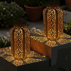 Könnte beinhalten: Zwei goldfarbene Solar-Laternen mit aufwendigen Ausschnitten, die Lichtmuster auf die Oberfläche werfen. Jede Laterne hat einen Griff. Der Hintergrund zeigt Grün und einen Blumentopf. Geeignet für den Außenbereich.