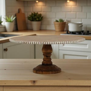 May include: A wooden cake stand with a round, light-brown top and a decorative white edge. The stand has a turned, dark-brown pedestal base and a matching round foot. The stand is displayed on a light-colored wooden surface in a kitchen setting.