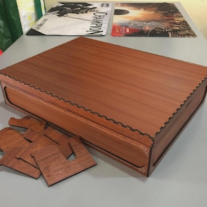 Könnte beinhalten: Eine rechteckige Aufbewahrungsbox aus braunem Holz mit flachem Deckel und lasergeschnittenen Kanten. Mehrere kleine, flache Holzteile mit Aussparungen liegen neben der Box, die eine glatte Oberfläche mit einer sichtbaren Holzmaserung aufweist.